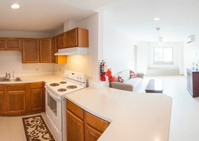 A view of a kitchen with wooden cabinets and white countertops. To the right you can see the white living room and a hallway