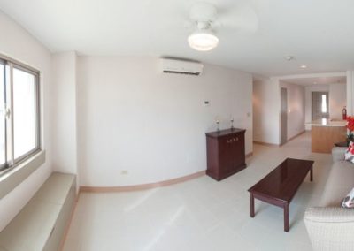 A view of a clean white living room. There is a window to the left. The living room contains a couch, coffee table, TV stand, and ceiling fan. To the right there is a hallway and an archway leading to the kitchen